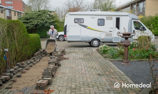 Man brengt zand aan met een kruiwagen op een opgebroken oprit, waar gebakken klinkers opnieuw worden aangelegd in een woonwijk met een camper op de achtergrond.