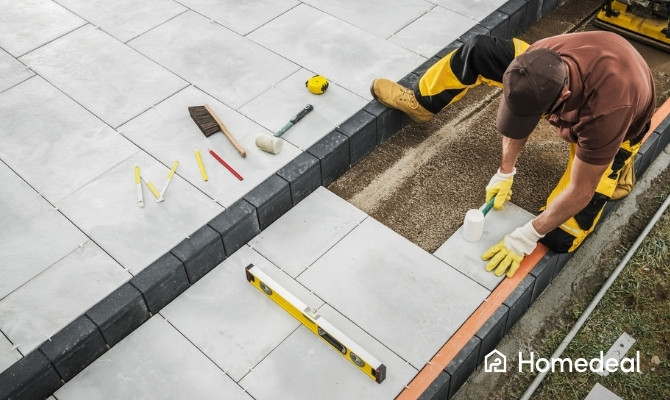 Stratenmaker plaatst grote grijze tuintegels op een stabilisé ondergrond, met behulp van rubberhamer, afstandhouders en waterpas; diverse gereedschappen liggen klaar op het terras.