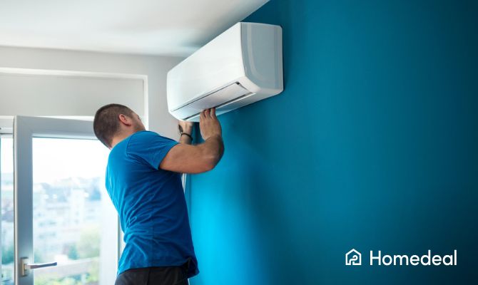 Monteur in blauwe kleding plaatst een wandmodel airco tegen een felblauwe muur in een moderne ruimte met daglicht.