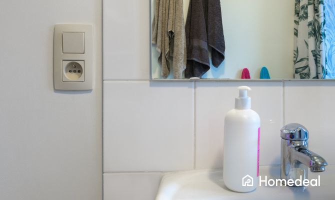 Stopcontact met een schakelaar in een badkamer - je ziet een stuk van een wastafel en spiegel naast het stopcontact
