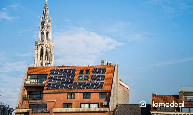 Modern appartementsgebouw met zonnepanelen op het schuine dak, met op de achtergrond de toren van de Onze-Lieve-Vrouwekathedraal in Antwerpen onder een blauwe hemel.