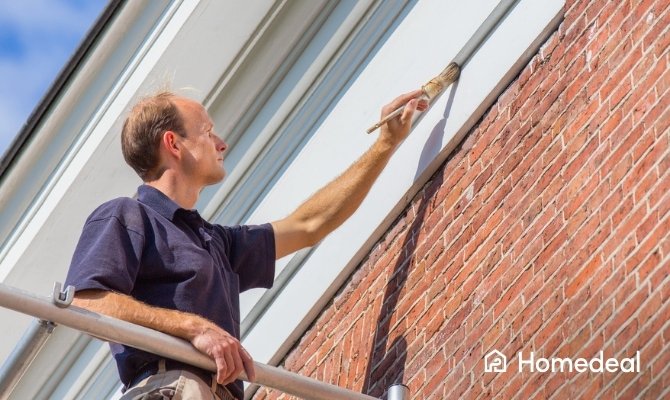 Buitenschilder schildert houten gevelrand wit boven een bakstenen muur terwijl hij op een steiger staat.