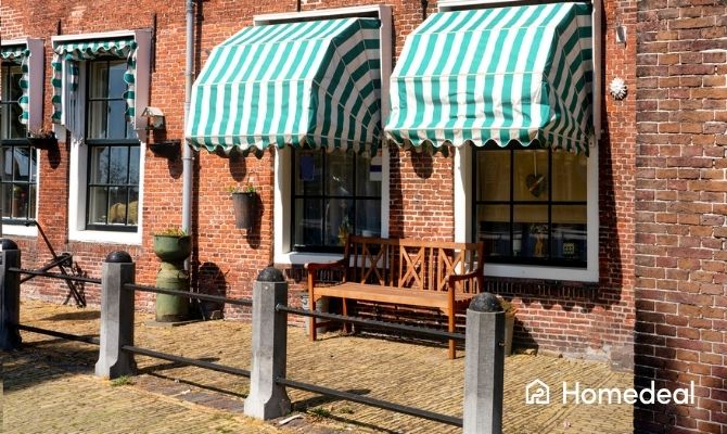 Traditionele markiezen met groen-wit gestreept doek aan een rood bakstenen gevel, boven ramen met zwarte kozijnen en een houten bank eronder.