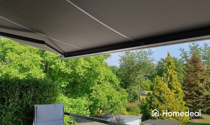 Grijs knikarmscherm boven een balkon met tuinstoel en uitzicht op groene bomen, als zonwering voor een terras.