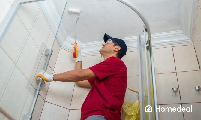 Binnenschilder schildert het plafond van een badkamer met een verfroller terwijl hij een douchecabine vasthoudt.
