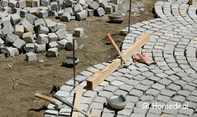Herbestraten van grijze kasseien in een gebogen patroon op een zandbed, met richtsnoeren, houten latten en bestratingsgereedschap op de werf.