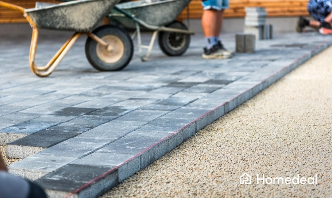 Werken aan tuinbestrating met grijze betonklinkers; tuintegels worden gelegd op een zandbed met behulp van een touwlijn voor een rechte plaatsing.