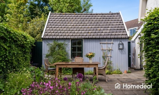 Tuinhuis met grijze dakbedekking van betonnen dakpannen in een groene tuin, met houten tuintafel, stoelen en bloeiende planten op het terras.