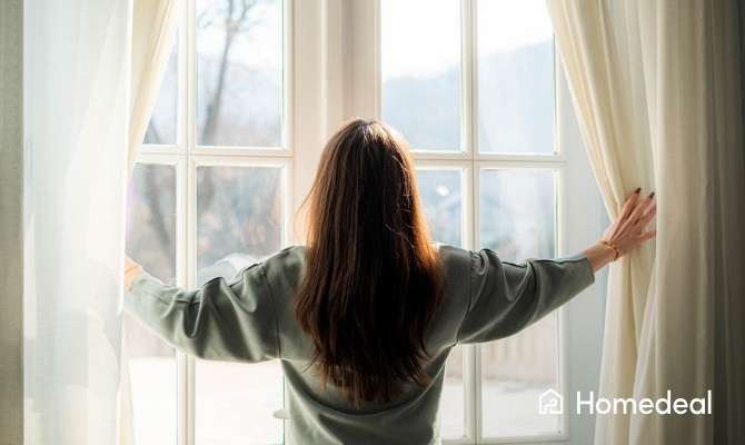 Persoon opent de gordijnen bij een groot vast raam en laat natuurlijk daglicht binnen in de woning.