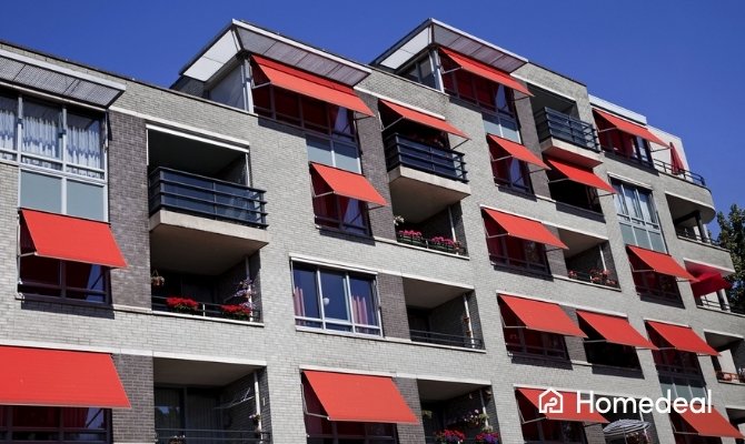 Appartementengebouw met meerdere rode uitvalschermen als zonwering boven ramen en balkons onder een heldere blauwe lucht.