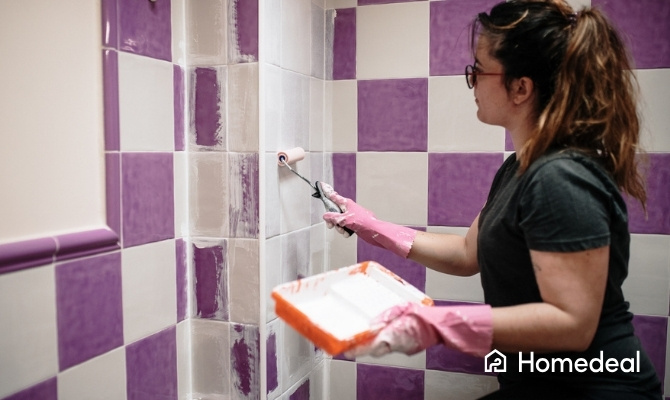 Vrouw met roze handschoenen schildert paarse tegels in de badkamer wit met een kleine verfroller en verfbakje.