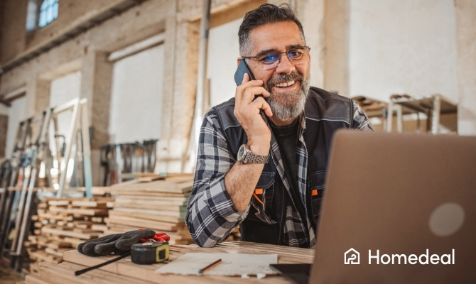 Schrijnwerker in de buurt die in zijn werkplaats telefoneert terwijl hij op een laptop offertes of plannen bekijkt, omringd door houten planken en gereedschap.