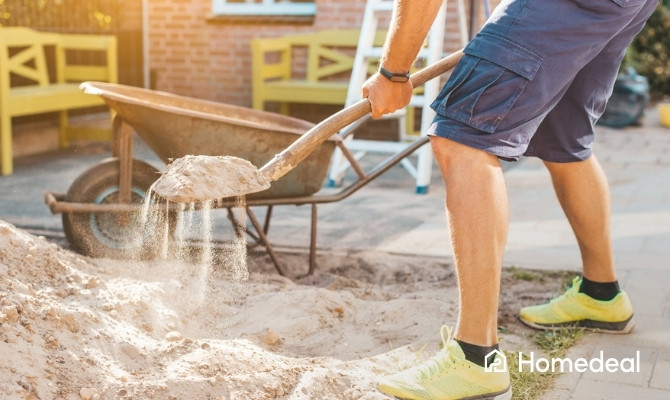 Persoon die zand egaliseert met een schop als voorbereiding van de fundering voor een tuinhuis in de tuin.