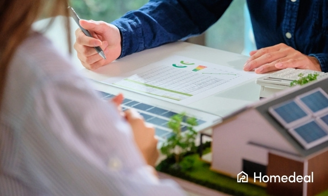 Twee personen bespreken een energieoverzicht aan tafel met een laptop en een maquette van een huis met zonnepanelen, tijdens het berekenen van hoeveel zonnepanelen nodig zijn.