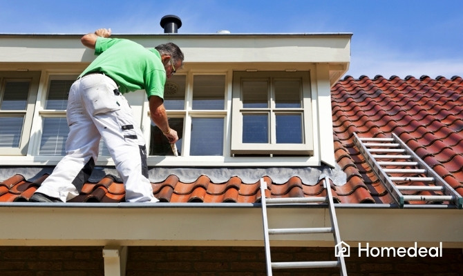 Schilder staat op een dak met rode dakpannen en is bezig met dakkapel schilderen. Hij verft de houten kozijnen van de dakkapel met een kwast, terwijl twee ladders tegen het dak staan.