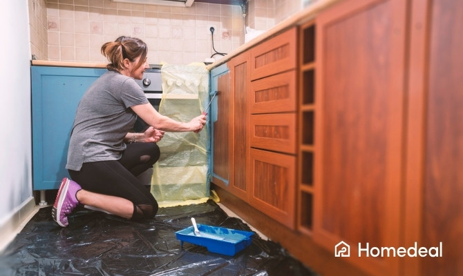 Vrouw zit op de grond en verft houten keuken blauw met een kwast. De vloer is afgedekt met plastic en er staat een verfrolbak met blauwe verf naast haar.