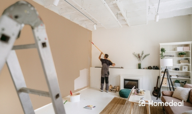 Schilder verft een muur in de woonkamer beige met een lange verfroller. Op de grond staan een verfemmer en kleurenstalen, terwijl meubels en decoratie zichtbaar zijn in de ruimte.
