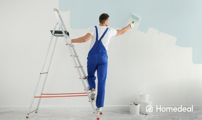 Schilder in blauwe overall staat op een trap en verft een witte muur lichtblauw met een verfroller. Op de vloer liggen afdekzeilen en verfemmers.