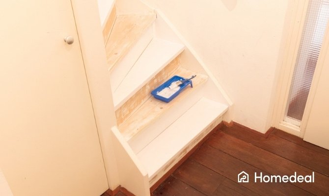 Houten trap in renovatie waarbij de treden deels geschuurd en deels wit zijn geverfd. Op de trap staat een blauwe verfrolbak met witte verf en een kwast, klaar voor verder trap schilderen.