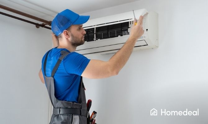 Airco installateur in blauwe werkkleding en pet voert onderhoud uit aan een wandgemonteerde airconditioning binnen in huis.
